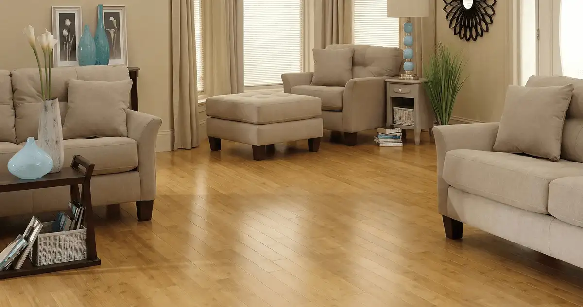 A living room with professionally installed bamboo flooring, beige sofas, a matching ottoman, a side table with a blue lamp, vases, framed pictures, and large windows with blinds and curtains in Albuquerque, NM